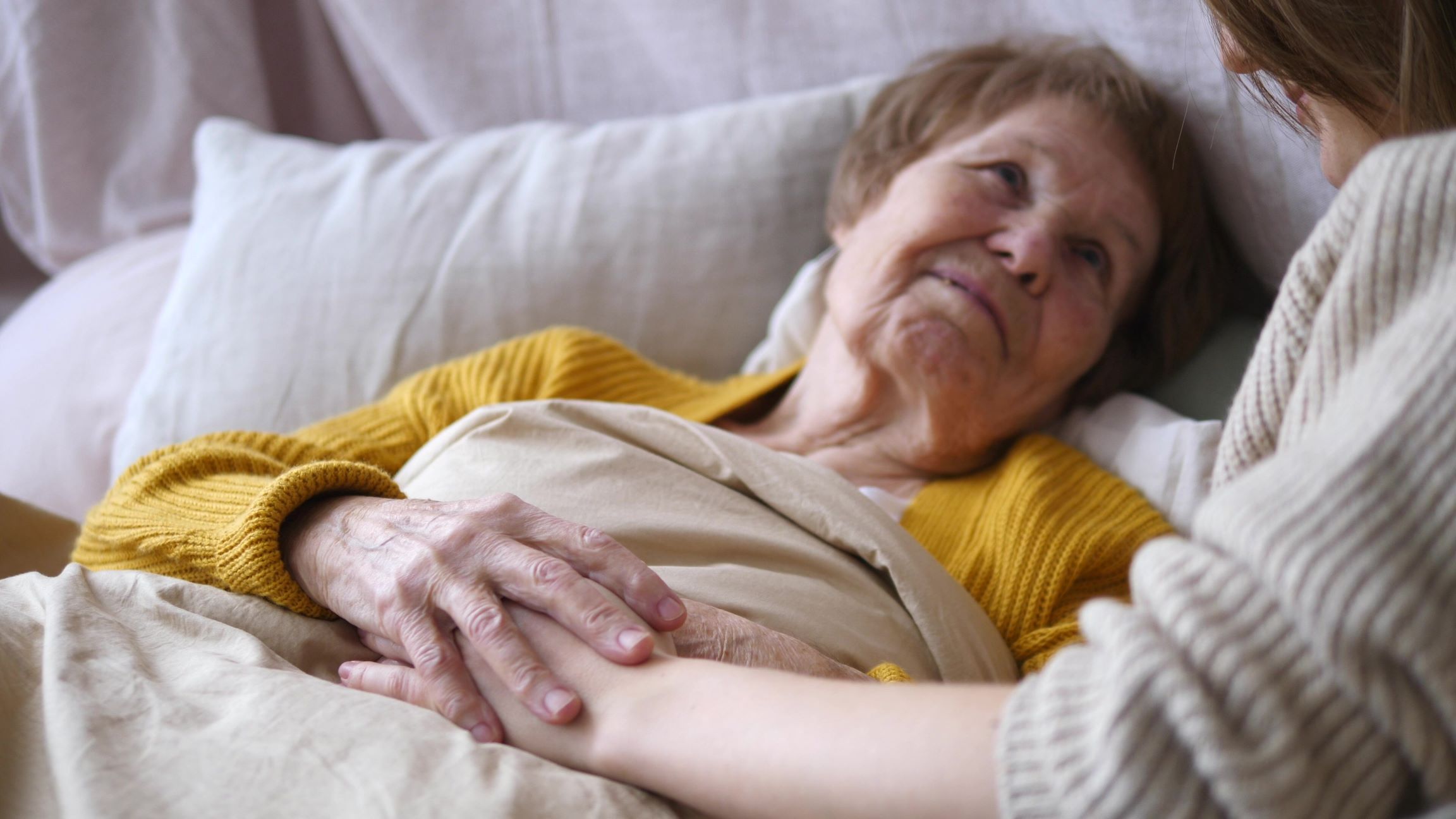 Close up of senior woman lying in bed as she looks up at her adult daughter caregiver.